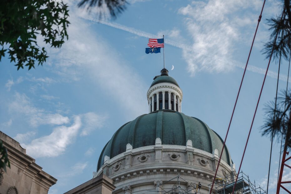 The Indiana State Capitol dome with American and Indiana flags flying against a bright blue sky.